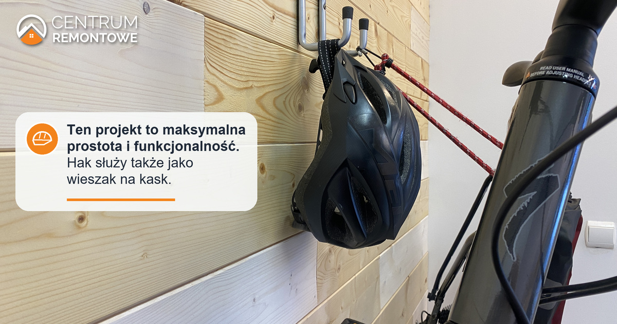 Rower na uchwycie umieszczony na drewnianej ściance. Jest odchylony lekko w bok, co pozwala zmieścić kierownicę i powiesić na haku dodatkowe akcesoria - na tym ujęciu jest to kask rowerowy.
